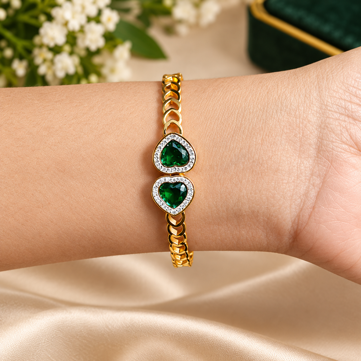 Close-up of a golden link bracelet featuring two emerald-green heart-shaped crystals surrounded by crystal halos, worn elegantly on a wrist.