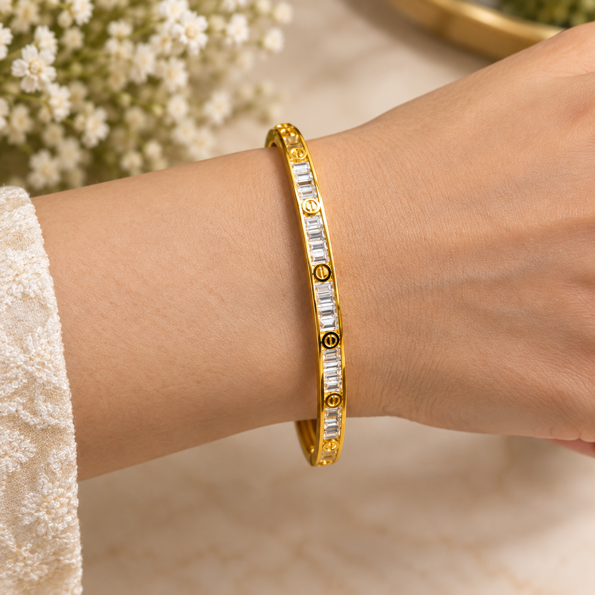 Close-up of a golden bangle bracelet featuring channel-set baguette crystals and screw-motif accents worn on a wrist.   