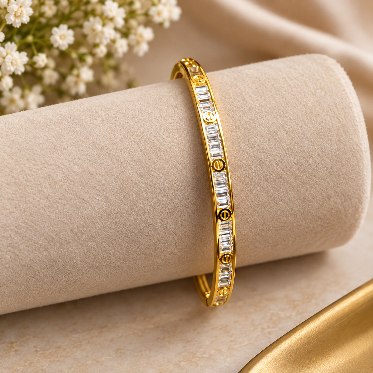 Studio product display of a gold-tone crystal baguette bangle resting on a beige padded stand.   