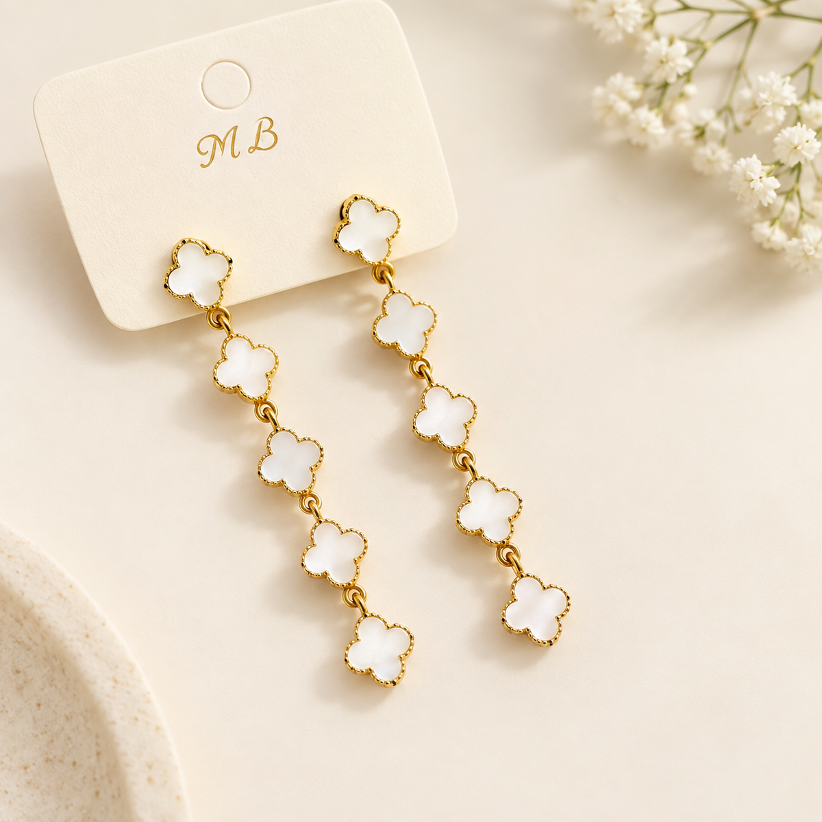 Studio display of golden drop earrings featuring cascading white clover motifs attached to a branded card on a neutral surface.   
